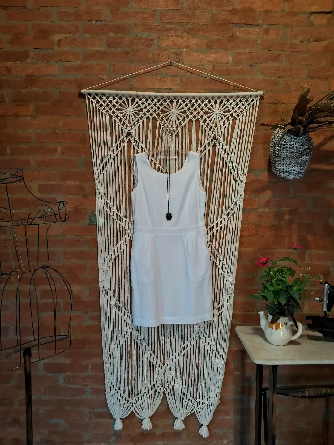 Vestido branco de linho pendurado em macramê sobre parede de tijolos
